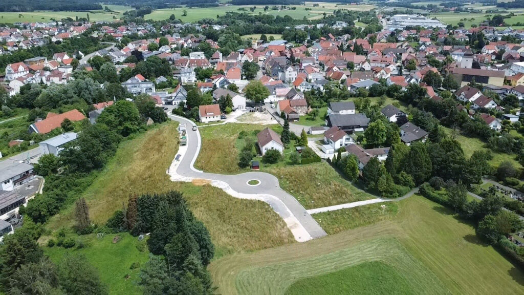 Luftaufnahme einer kleinen Stadt mit Häusern, Bäumen und Gärten. Ein Kreisverkehr befindet sich am Ende einer teilweise ausgebauten Straße bei Kreisbau Ostalb, umgeben von grünen Feldern und unbebauten Flächen.