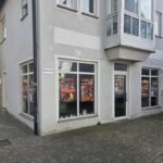 Ein Eckhaus mit großen Fenstern, an denen bunte Plakate hängen, die wahrscheinlich für Lebensmittel werben. Die Straße ist mit Kopfsteinpflaster gepflastert, und ein Schild an der Wand lautet Spitalstraße 23. Einige Pflanzen stehen in der Nähe des Eingangs.