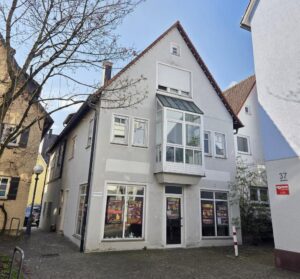 Ein zweistöckiges, hellgraues Gebäude mit großen Fenstern an der Vorderseite, auf denen Plakate angebracht sind, einem kleinen überdachten Balkon im Obergeschoss und einem blattlosen Baum davor, an einer Kopfsteinpflasterstraße unter freiem Himmel gelegen.