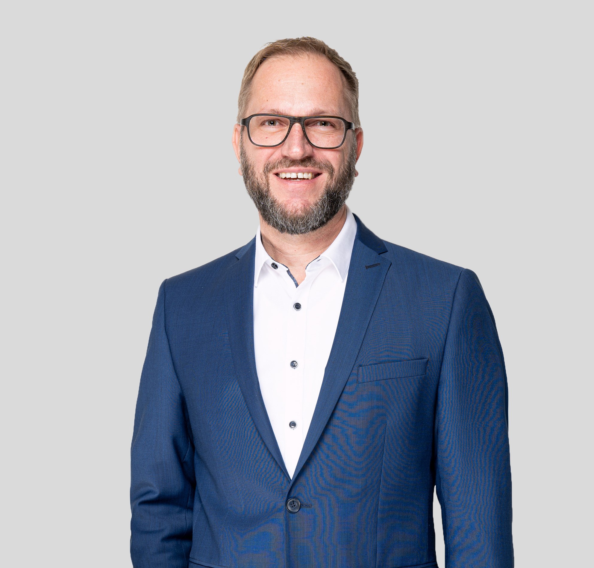 Ein lächelnder Mann mit Brille, Bart und kurzen Haaren trägt ein blaues Sakko über einem weißen Hemd und steht vor einem schlichten hellgrauen Hintergrund.
