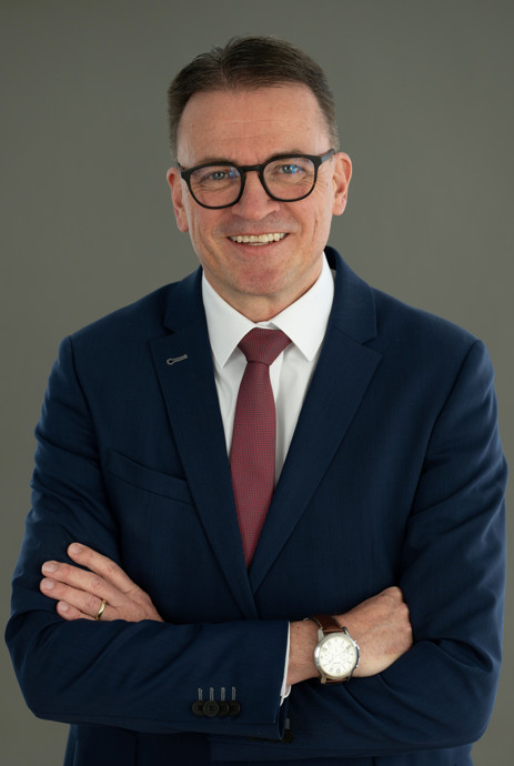 Ein Mann in einem marineblauen Anzug, einem weißen Hemd und einer roten Krawatte steht mit verschränkten Armen da und lächelt. Er trägt eine Brille und eine Armbanduhr und hat kurzes braunes Haar. Der Hintergrund ist schlicht und grau.